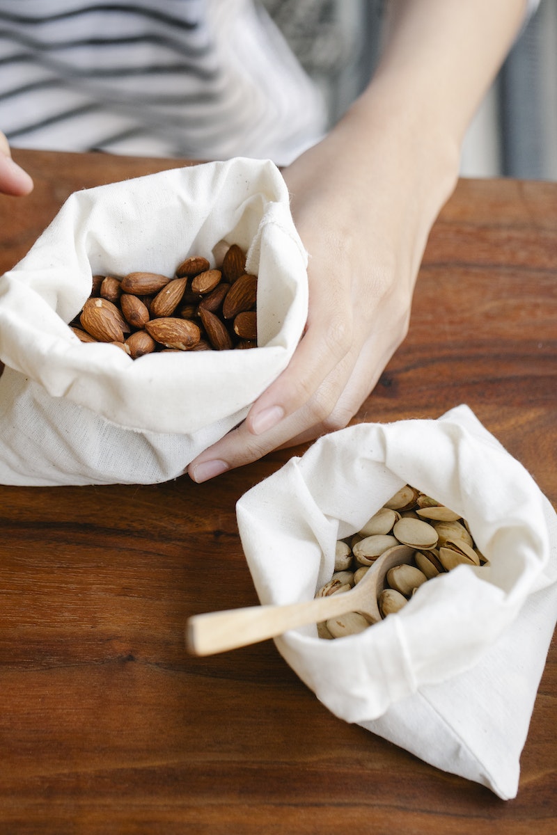 Crop person showing almonds in white bag near pack of pistachios with wooden spoon on table